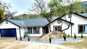 A two-story house with a garage, a driveway, and a landscaped area with a rock feature and small plants. - Oasis Riverfront House - Gazebo overlooks the River, 5 miles to Sequoia SNP gate (Three Rivers)