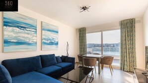 Een moderne woonkamer met een blauwe bank, een glazen salontafel en een eethoek met stoelen. Twee grote schilderijen hangen aan de muur en er is uitzicht op de stad door grote ramen.
