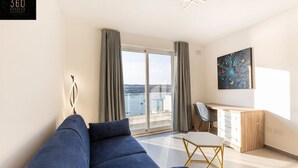 Een moderne woonkamer met een blauwe bank, een bureau en een stoel. Er is een groot raam met gordijnen, een schilderij aan de muur en uitzicht op zee.