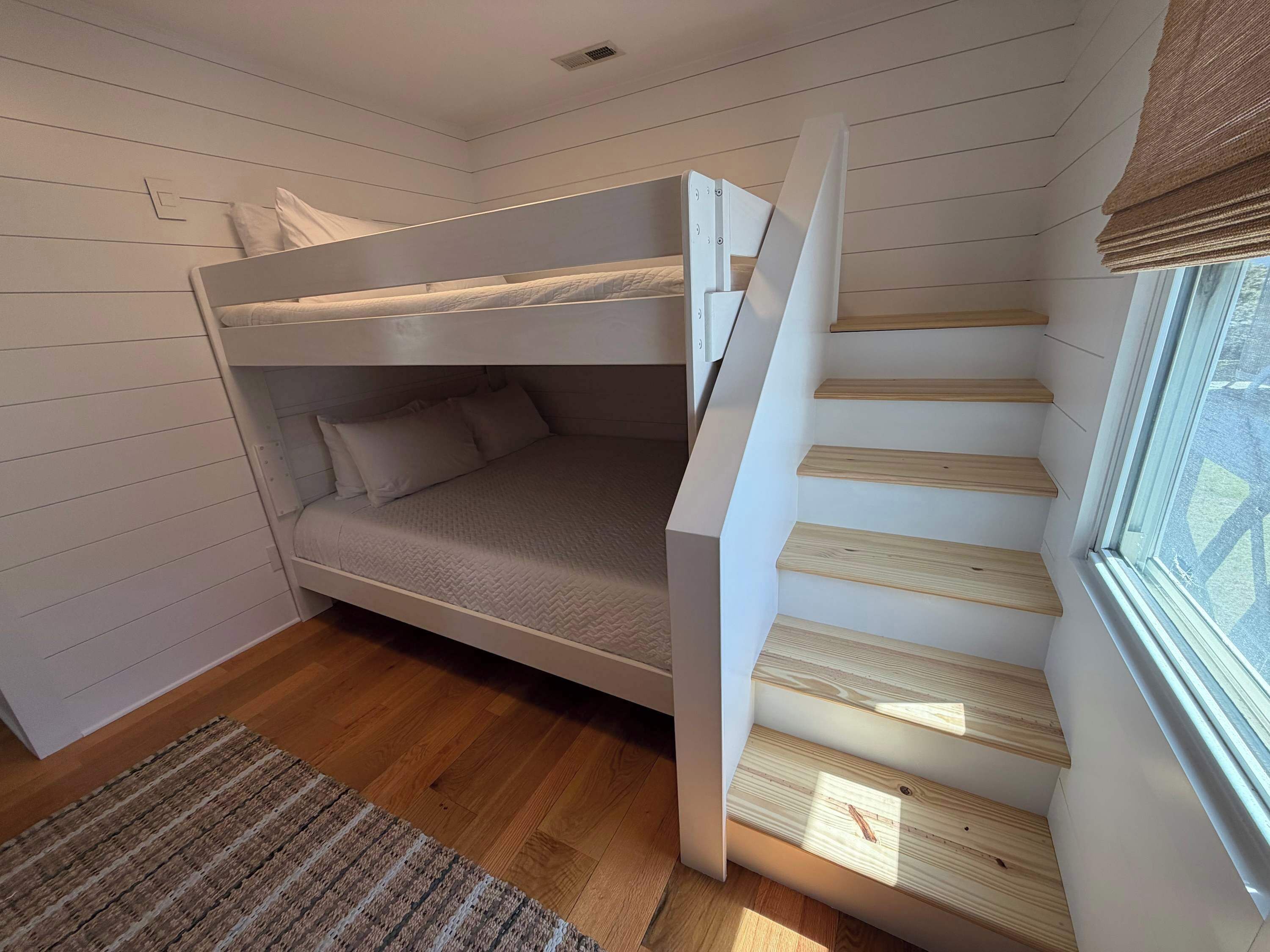 Un lit superposé dans une pièce dont le sol est recouvert de planches de bois, avec une fenêtre équipée de stores et un petit tapis.