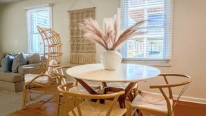A room with a round table, wicker chairs, a white vase with pampas grass, a woven wall hanging, and a sofa with cushions.