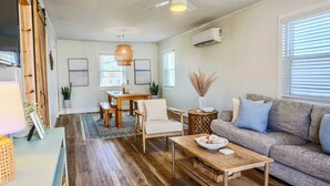 A living room with a wooden floor, a sofa, a coffee table, a dining table with chairs, and a hanging light fixture.