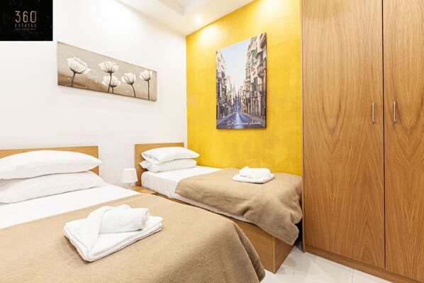 A hotel room with two beds, a wooden wardrobe, a painting of a street, and a floral artwork on the wall. - Seafront 4BR APT with mesmerising Balcony & VIEWS (Tas-Sliema)