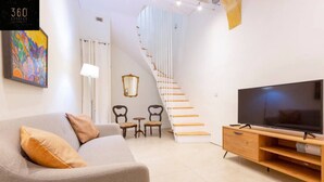 Een moderne woonkamer met een grijze bank, een televisie op een houten dressoir, en een trap naar een ander niveau.