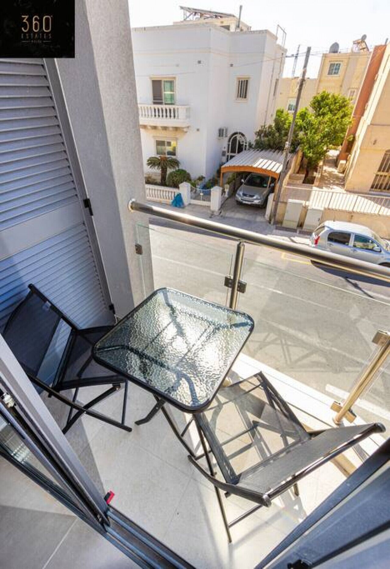 A balcony with a glass table and chairs, a metal railing, and a view of a street with buildings and parked cars.