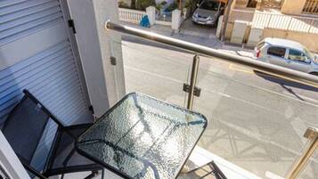 A balcony with a glass table and chairs, a metal railing, and a view of a street with buildings and parked cars.