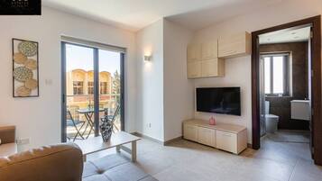 A modern living room with a sofa, a TV on a wooden cabinet, and a balcony with a view of buildings.