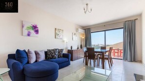 A modern living room with a blue sofa, a glass coffee table, and a dining area with a table and chairs. There is a large window with curtains, a chandelier, and artwork on the walls. - Sliema Ferries APT, with beautiful views AC & WIFI (Sliema)