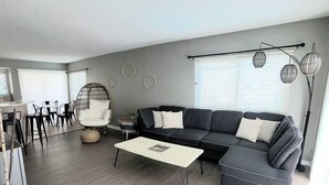 A modern living room with a sectional sofa, a dining area with chairs, a hanging chair, and a floor lamp. - Oceanfront Lookout, pet friendly with pool (Carolina Beach)