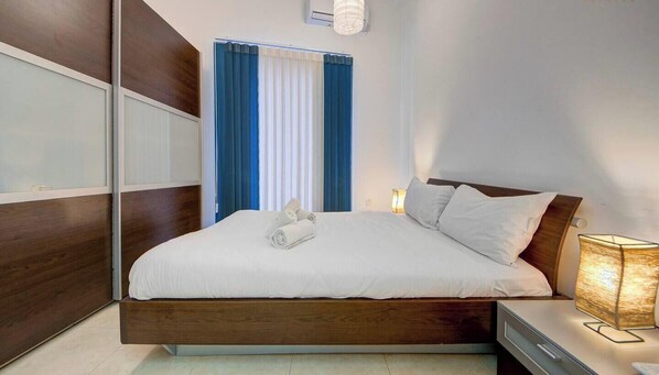 A modern bedroom with a wooden bed frame, a bedside lamp, and a window with blue curtains. - Central APT 5mins to promenade with SOFA AC & WIFI (Sliema)