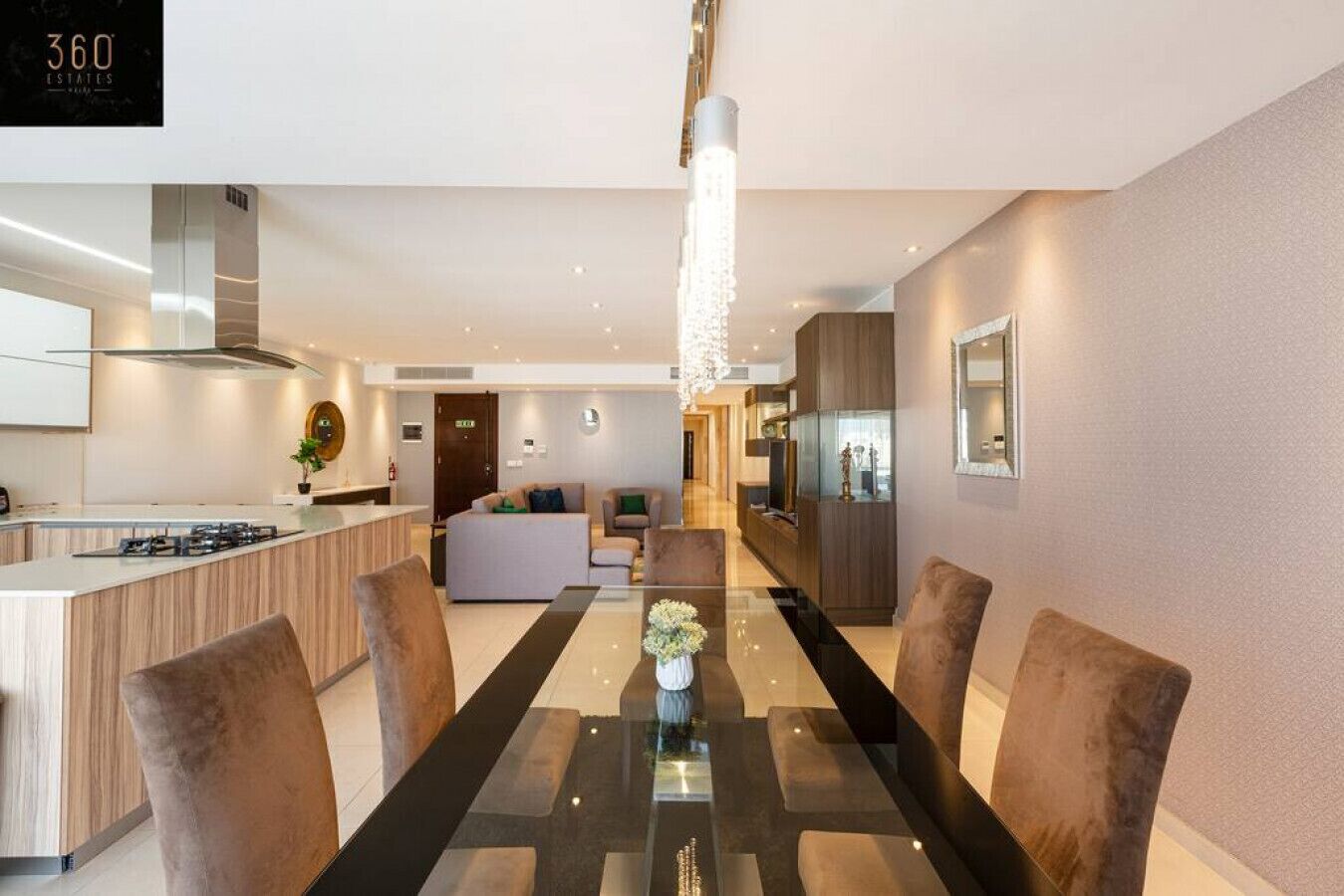 Una cocina moderna con una isla central, un área de comedor con una mesa de vidrio y un espacio de sala de estar con sofá y televisor.