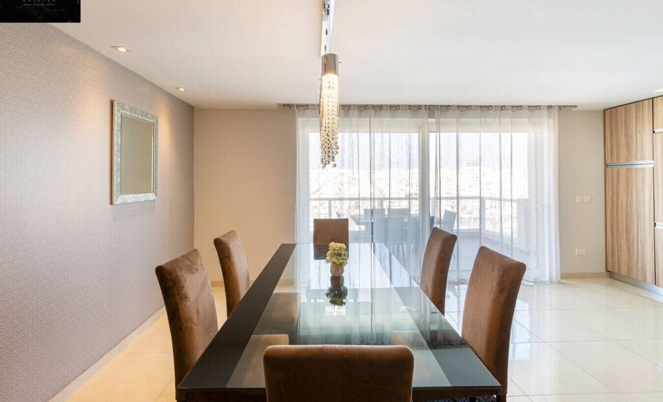 Een moderne eetruimte met een glazen tafel, beklede bruine stoelen, een hanglamp en uitzicht door grote ramen.
