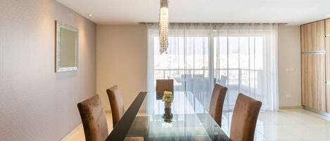 Een moderne eetruimte met een glazen tafel, beklede bruine stoelen, een hanglamp en uitzicht door grote ramen.