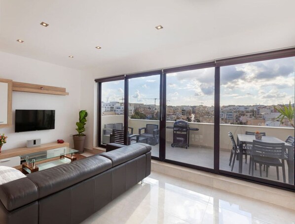 A modern living room with a large window, a flat-screen TV, a sofa, and a balcony with outdoor furniture. - Penthouse + BBQ Kitchen Island & Massive living (San Ġiljan)