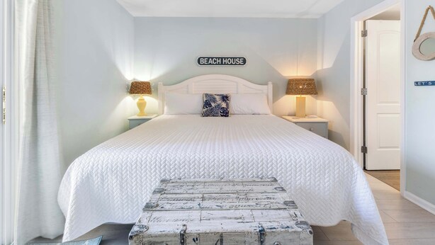 A bedroom with a large bed, two bedside tables, a headboard sign reading 'BEACH HOUSE', and a wooden chest. - ★ 3 Little Birds at The Beach★ (Carolina Beach)