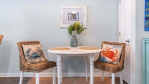 A dining area with two wicker chairs, a white table, and a framed picture on the wall. - ★ 3 Little Birds at The Beach★ (Carolina Beach)