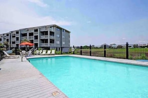 A swimming pool with a wooden deck, lounge chairs, and a building with balconies in the background. - ★ 3 Little Birds at The Beach★ (Carolina Beach)