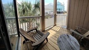 A wooden deck with a bench and a table, a glass door, and a view of palm trees and buildings. - The Narrow Nest @ Sand Pebbles (Carolina Beach)