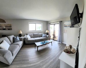 A spacious living room with a grey sofa, a coffee table, and a TV mounted on the wall.