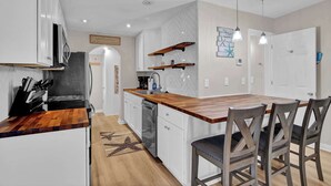 A modern kitchen with white cabinets, a wooden countertop, and a built-in dishwasher. There are two pendant lights, a wooden shelf, and a sign on the wall. - Beachwood Bungalow B, walk to beach and boardwalk (Carolina Beach)