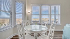 A dining area with a round glass table surrounded by white chairs, large windows with blinds, and a view of the beach. - Island Treasure, CB Oceanfront Retreat (Carolina Beach)