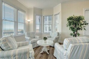A room with a round glass table surrounded by striped armchairs, large windows with blinds, and a potted plant.