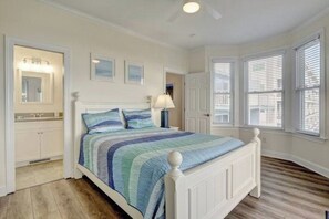 A bedroom with a bed, a nightstand, a lamp, a window with blinds, and a door. - Island Treasure, CB Oceanfront Retreat (Carolina Beach)