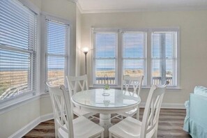 A dining area with a round glass table and white chairs, large windows with blinds, and a view of the beach. - Island Treasure, CB Oceanfront Retreat (Carolina Beach)