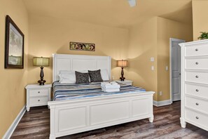 A bedroom with a white bed, two nightstands with lamps, a dresser, and a door leading to another room.