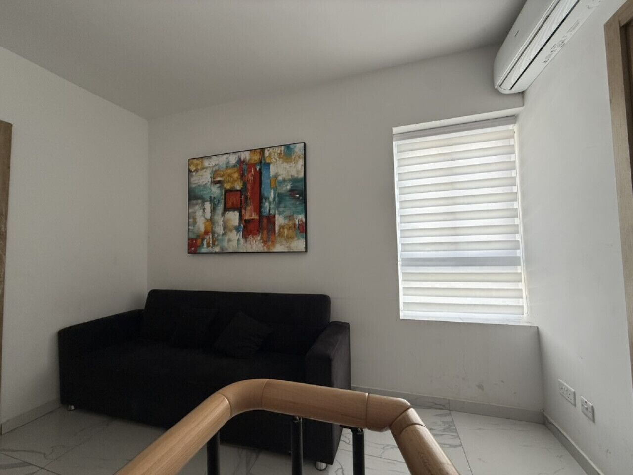 A living room with a black sofa, a colorful abstract painting, a window with blinds, and a wooden chair.