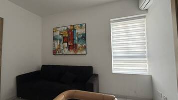 A living room with a black sofa, a colorful abstract painting, a window with blinds, and a wooden chair.
