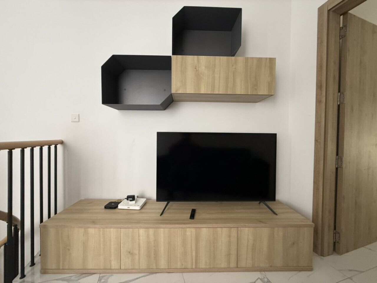 A modern living room with a wooden TV stand, a flat-screen TV, and three black shelves.