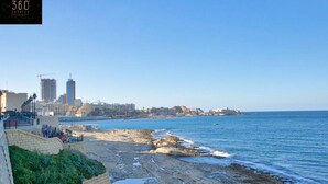 A coastal cityscape with a rocky beach, a promenade, and a clear view of the sea. - Sliema Seafront 3BR - opposite Beach - AC & Wifi (Sliema)