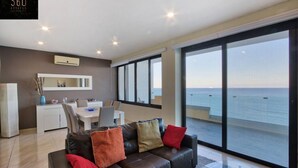 A modern living room with a leather sofa, a dining area, and large windows offering a view of the sea. - Sliema Seafront 3BR - opposite Beach - AC & Wifi (Sliema)