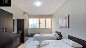 A hotel room with two beds, a desk, a wardrobe, and a window with a city view. - Sliema Seafront 3BR - opposite Beach - AC & Wifi (Sliema)