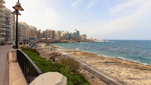 A coastal promenade with a stone pathway, a stone lantern, and a view of the sea and buildings in the background. - Spacious Bright Seafront APT (Tas-Sliema)
