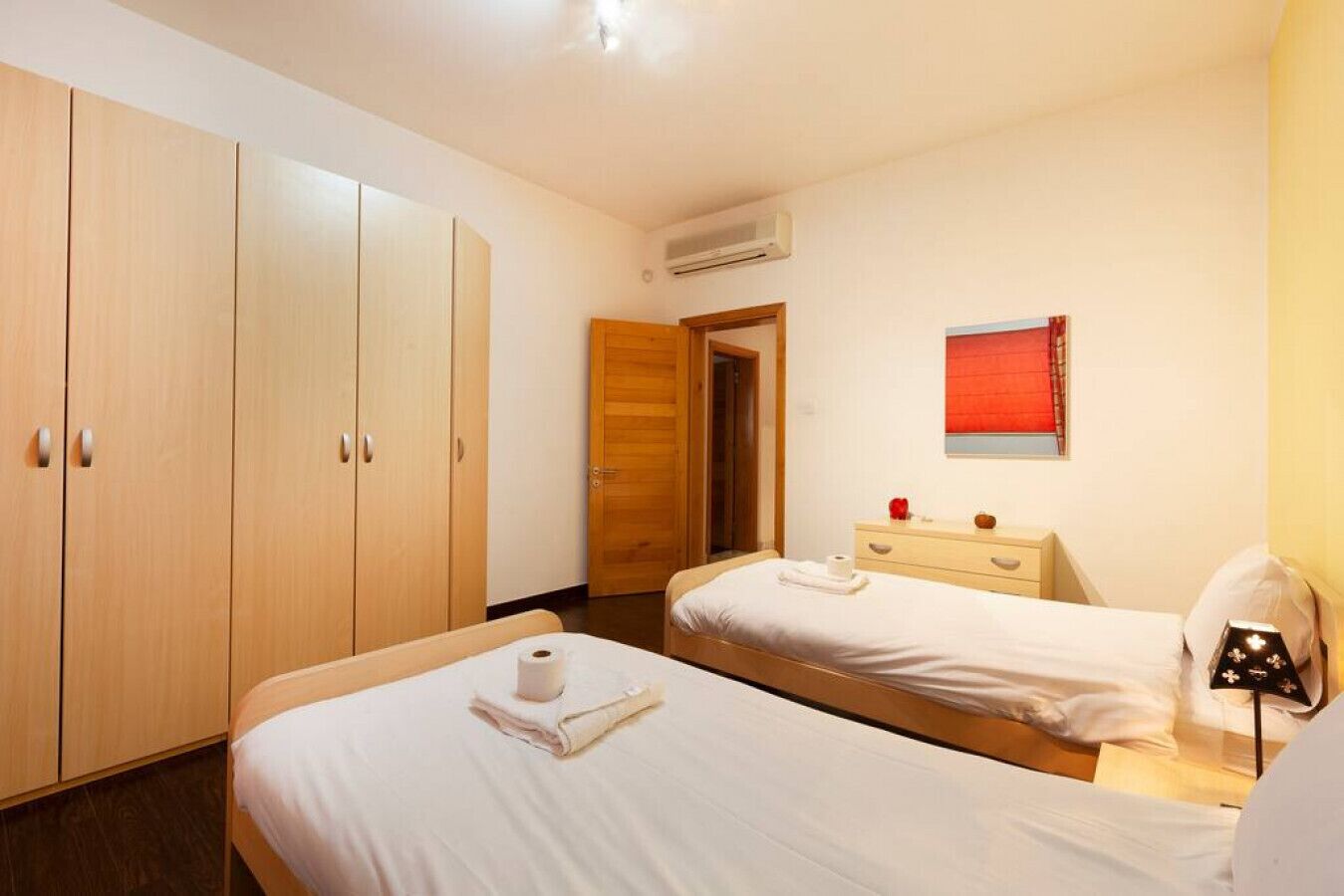 Une chambre d’hôtel avec deux lits, une armoire en bois, une table de chevet avec une lampe et une fenêtre avec un store rouge.
