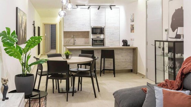 A modern kitchen with white cabinets, a central island, and a dining area with a round table and chairs. - Beautiful, cozy APT just off the Promenade & WIFI (Tas-Sliema)
