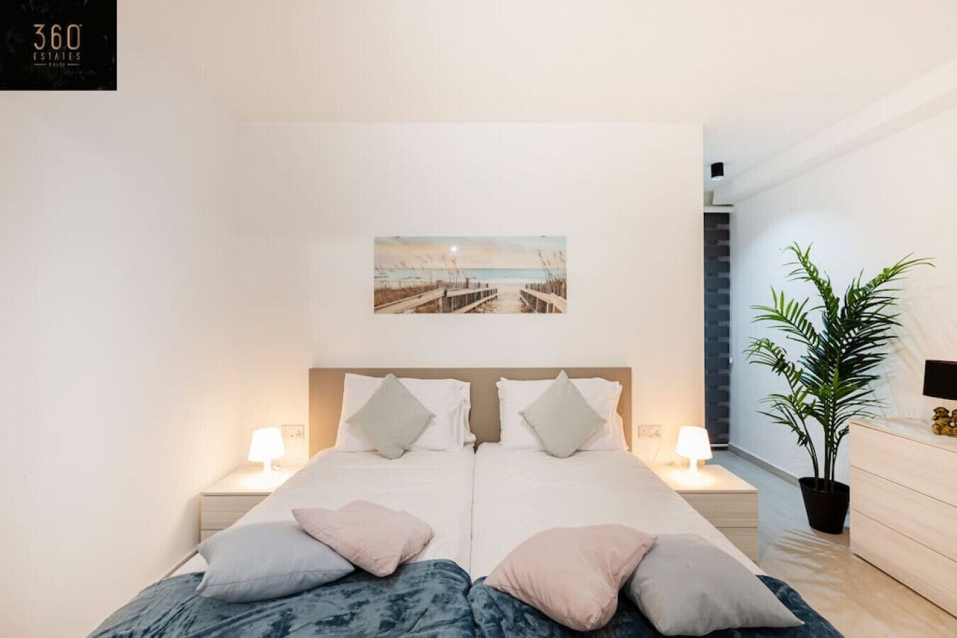 Une chambre moderne avec un grand lit, deux tables de chevet avec des lampes, une plante en pot et une œuvre d’art sur le thème de la plage accrochée au mur.