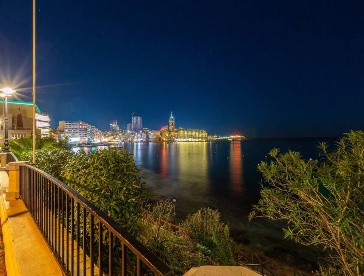 A waterfront promenade with a city skyline in the background, featuring illuminated buildings and a calm sea.