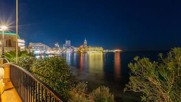 A waterfront promenade with a city skyline in the background, featuring illuminated buildings and a calm sea.