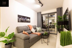 A modern living room with a grey sofa, a cityscape painting, and a view into the kitchen area. - Beautiful, cozy APT just off the Promenade & WIFI (Tas-Sliema)