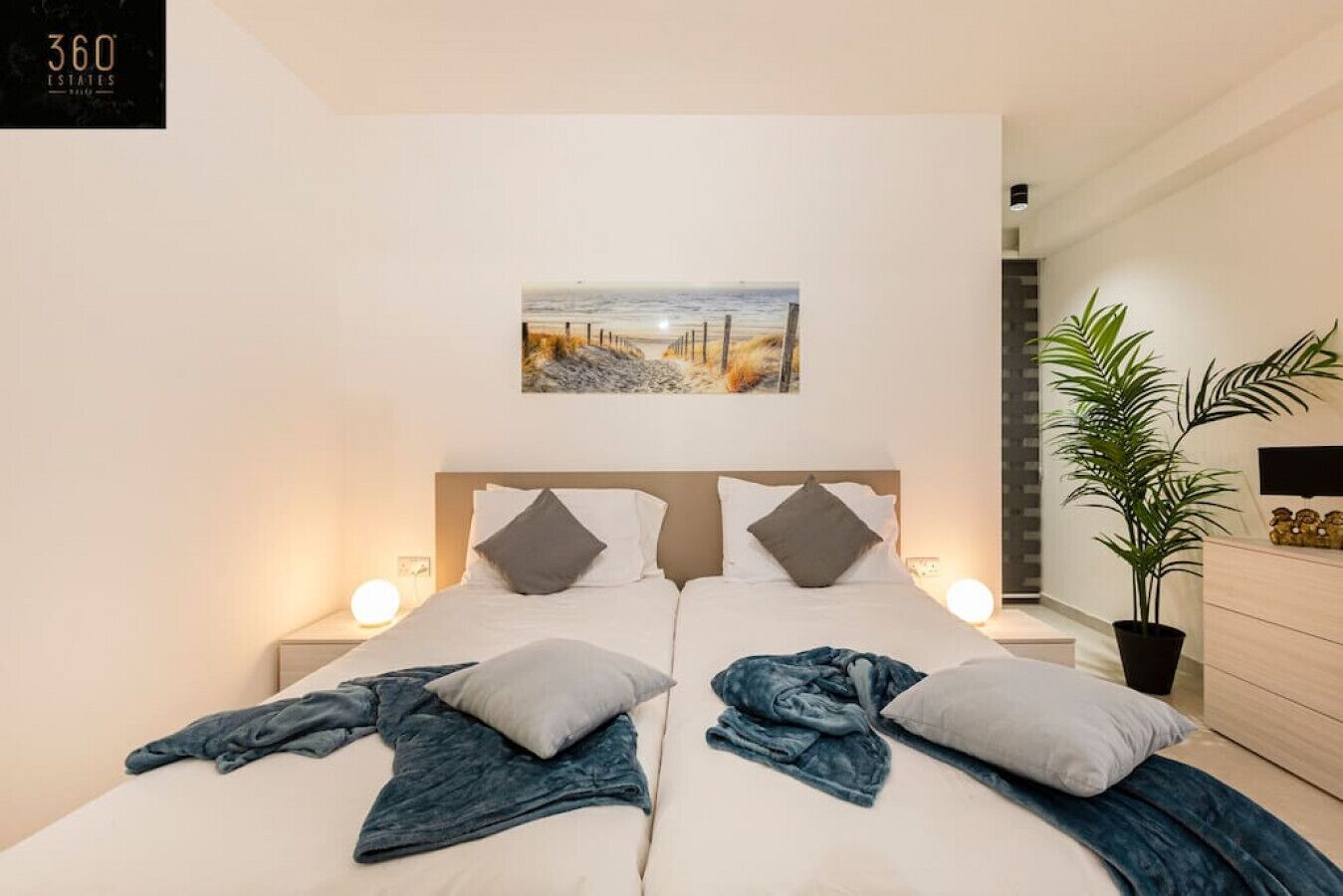 A hotel room with two beds, a painting above the beds, a potted plant, and a sign on the wall.