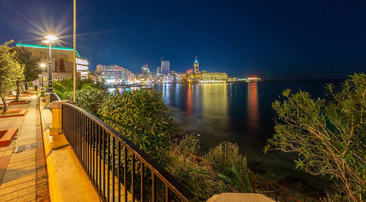 A waterfront promenade at night with a city skyline in the background, a well-lit pathway, and a railing overlooking the water.