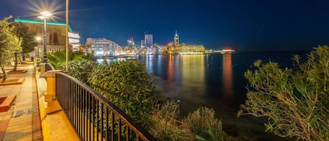 A waterfront promenade at night with a city skyline in the background, a well-lit pathway, and a railing overlooking the water.