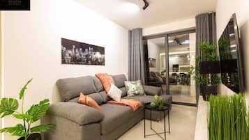 Un salon moderne avec un canapé gris, un tableau représentant un paysage urbain, une petite table sur laquelle se trouve un cactus et une plante en pot.