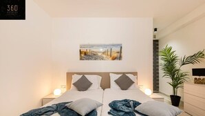 Une chambre moderne avec deux lits, un tableau représentant une scène de plage, des lampes de chevet, une plante en pot et une commode en bois.