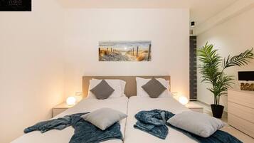 Une chambre moderne avec deux lits, un tableau représentant une scène de plage, des lampes de chevet, une plante en pot et une commode en bois.