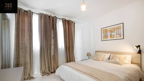 A bedroom with a large bed, a desk, and a chair. There is a window with curtains, a lamp, and a framed picture on the wall. - A beautiful, spacious penthouse (Il-Gżira)