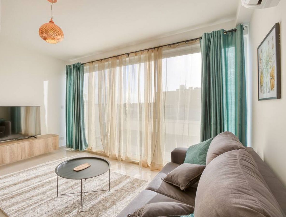 A modern living room with a flat-screen TV, a round coffee table, a sofa, and a large window with sheer curtains.